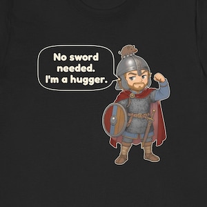 May include: A black t-shirt featuring a cartoon illustration of a warrior in chainmail and a red cape, wearing a helmet with a small boar on top. The warrior holds a round shield and flexes an arm. A speech bubble contains the text: No sword needed. I'm a hugger.
