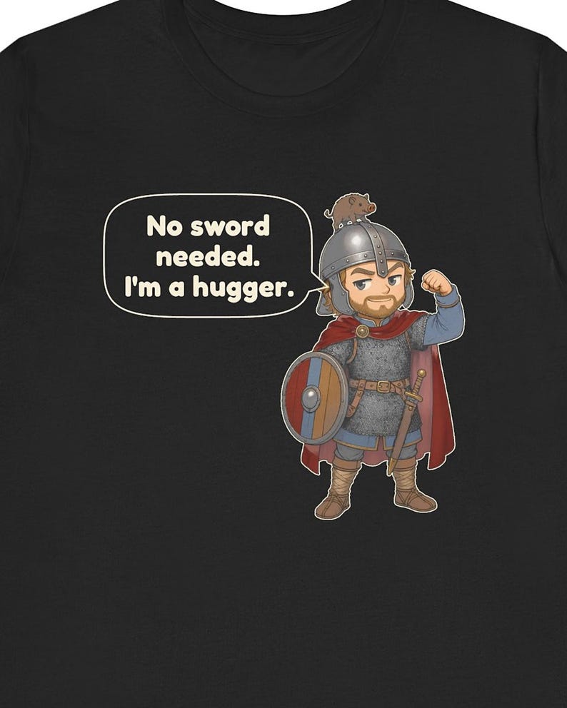 May include: A black t-shirt featuring a cartoon illustration of a warrior in chainmail and a red cape, wearing a helmet with a small boar on top. The warrior holds a round shield and flexes an arm. A speech bubble contains the text: No sword needed. I'm a hugger.