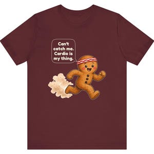 May include: A maroon t-shirt featuring a graphic of a running gingerbread person wearing a red and white headband. A speech bubble above the character contains the text: Can't catch me. Cardio is my thing. A small cloud of dust is behind the figure.