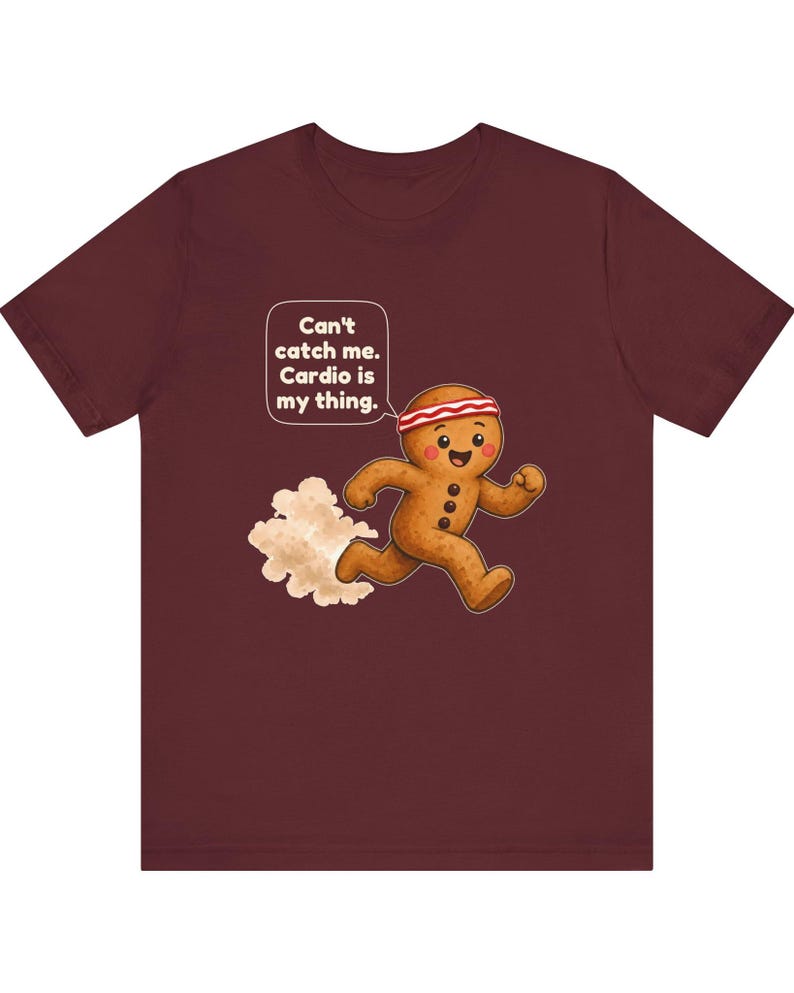 May include: A maroon t-shirt featuring a graphic of a running gingerbread person wearing a red and white headband. A speech bubble above the character contains the text: Can't catch me. Cardio is my thing. A small cloud of dust is behind the figure.