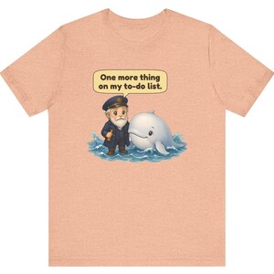 May include: A peach-colored t-shirt featuring a graphic of a bearded captain in a navy uniform standing next to a white whale in blue water. Above them, a speech bubble contains the text: One more thing on my to-do list.