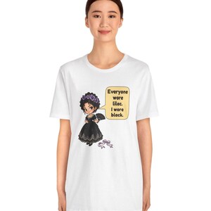 May include: A white t-shirt featuring a graphic of a character in a black dress with a purple flower crown and a fan. A speech bubble contains the text: Everyone wore lilac. I wore black. A small purple ribbon lies below the character.
