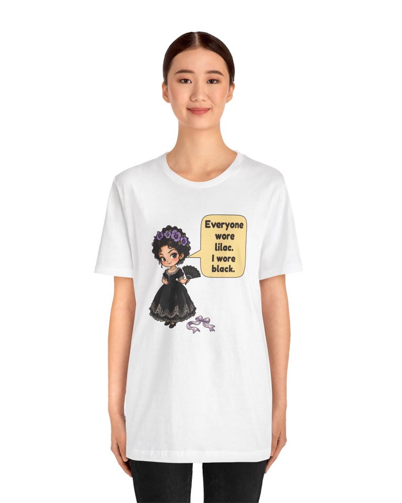 May include: A white t-shirt featuring a graphic of a character in a black dress with a purple flower crown and a fan. A speech bubble contains the text: Everyone wore lilac. I wore black. A small purple ribbon lies below the character.