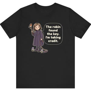 May include: A black t-shirt featuring a cartoon illustration of a person in a dark coat holding a key with a robin perched on it. A speech bubble contains the text: The robin found the key. I'm taking credit. Small purple flowers are visible.
