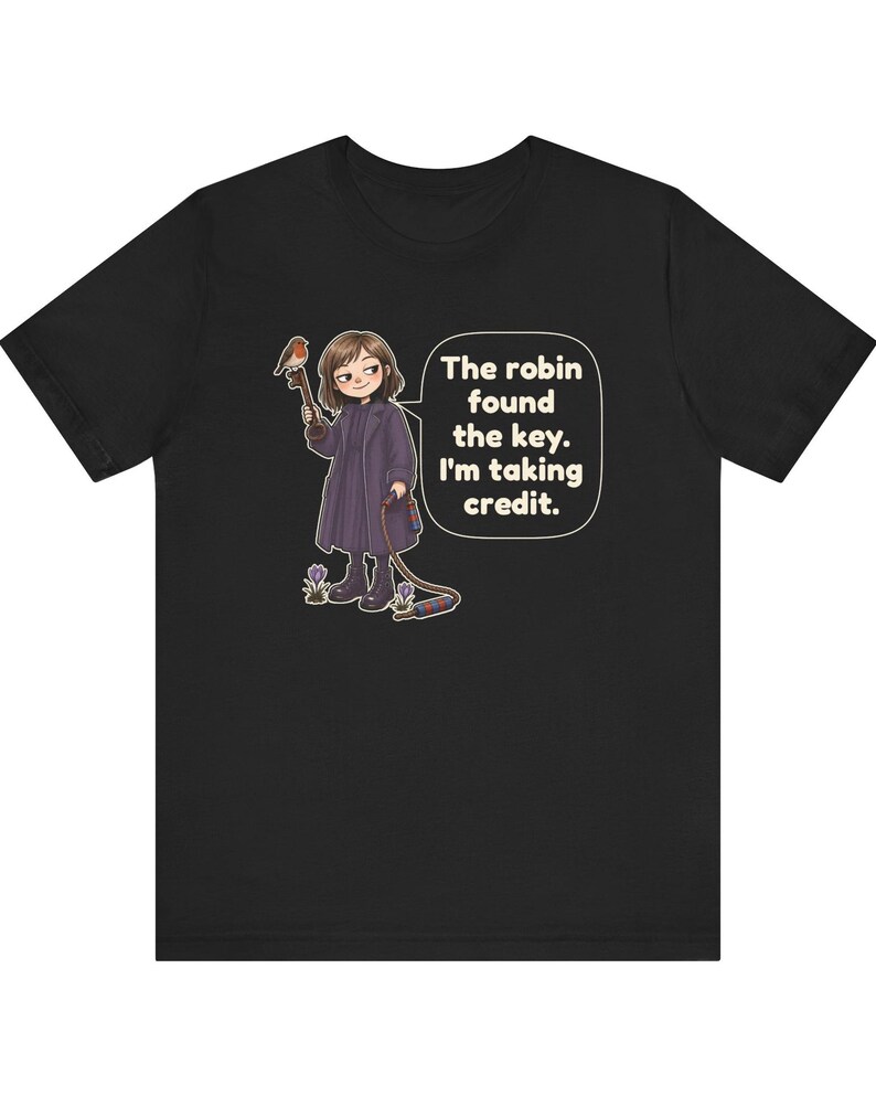 May include: A black t-shirt featuring a cartoon illustration of a person in a dark coat holding a key with a robin perched on it. A speech bubble contains the text: The robin found the key. I'm taking credit. Small purple flowers are visible.