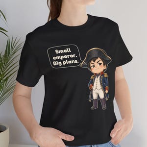 May include: A black short-sleeved t-shirt featuring a cartoon illustration of a historical figure in a blue military uniform and bicorne hat. A speech bubble next to the character contains the text: Small emperor. Big plans.