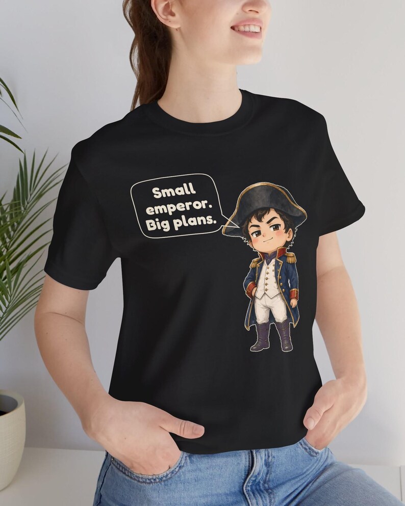 May include: A black short-sleeved t-shirt featuring a cartoon illustration of a historical figure in a blue military uniform and bicorne hat. A speech bubble next to the character contains the text: Small emperor. Big plans.