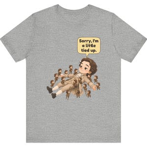 Op de afbeelding: Een grijs gem&ecirc;leerd t-shirt met een afbeelding van een groot figuur dat ligt, omringd door vele kleine figuurtjes die ladders en touwen gebruiken om het figuur vast te binden. Een tekstballon bevat de tekst, Sorry, I'm a little tied up.