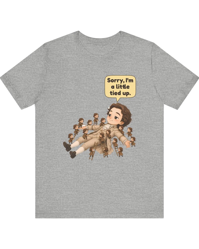 Op de afbeelding: Een grijs gem&ecirc;leerd t-shirt met een afbeelding van een groot figuur dat ligt, omringd door vele kleine figuurtjes die ladders en touwen gebruiken om het figuur vast te binden. Een tekstballon bevat de tekst, Sorry, I'm a little tied up.