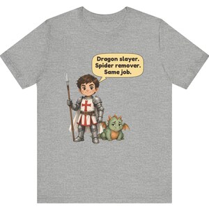 K&ouml;nnte beinhalten: Ein graumeliertes T-Shirt mit einer Cartoon-Illustration eines Ritters in silberner R&uuml;stung, der einen Speer h&auml;lt und neben einem kleinen gr&uuml;nen Drachen steht. Eine Sprechblase &uuml;ber dem Ritter enth&auml;lt den Text: Dragon slayer. Spider remover. Same job.