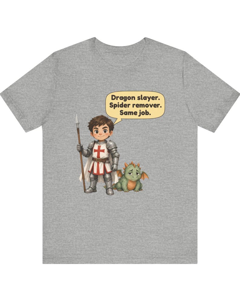 K&ouml;nnte beinhalten: Ein graumeliertes T-Shirt mit einer Cartoon-Illustration eines Ritters in silberner R&uuml;stung, der einen Speer h&auml;lt und neben einem kleinen gr&uuml;nen Drachen steht. Eine Sprechblase &uuml;ber dem Ritter enth&auml;lt den Text: Dragon slayer. Spider remover. Same job.