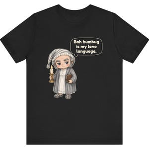 Puede incluir: Una camiseta negra con una ilustraci&oacute;n de dibujos animados de un personaje anciano con gorro de dormir y bata, sosteniendo una vela y un libro. Un globo de texto sobre el personaje contiene: "Bah humbug is my love language."