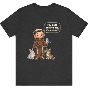 May include: A dark gray short-sleeve t-shirt featuring a cartoon illustration of a monk in a brown robe with a rabbit, a wolf, and a blue bird. A speech bubble above the monk contains the text: My pets talk to me. Yours too?