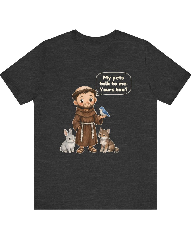 May include: A dark gray short-sleeve t-shirt featuring a cartoon illustration of a monk in a brown robe with a rabbit, a wolf, and a blue bird. A speech bubble above the monk contains the text: My pets talk to me. Yours too?