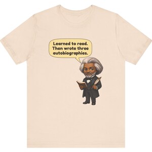 Puede incluir: Una camiseta de color beige claro con una ilustraci&oacute;n de dibujos animados de Frederick Douglass sosteniendo un libro y una pluma. Un globo de texto sobre la figura contiene el texto: Learned to read. Then wrote three autobiographies.