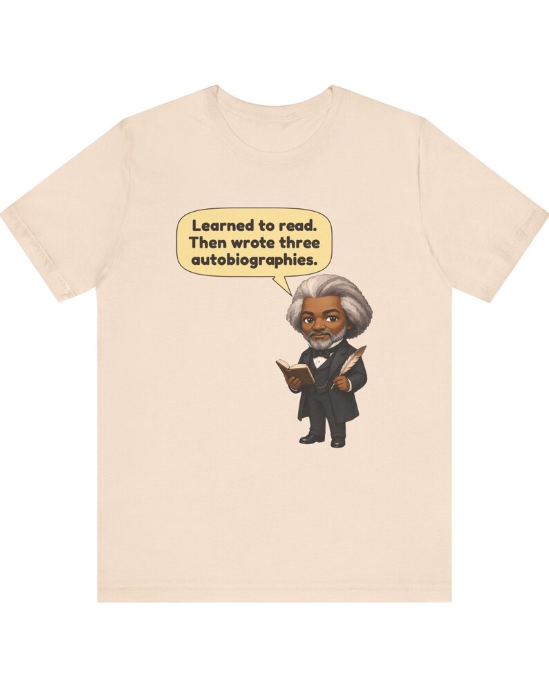 Puede incluir: Una camiseta de color beige claro con una ilustraci&oacute;n de dibujos animados de Frederick Douglass sosteniendo un libro y una pluma. Un globo de texto sobre la figura contiene el texto: Learned to read. Then wrote three autobiographies.