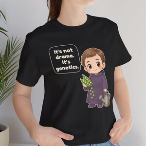 May include: A black t-shirt featuring a cartoon illustration of a monk holding a pea pod and a small watering can. A speech bubble next to the character contains the white text: It's not drama. It's genetics. The design is centered on the chest.