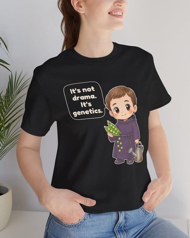 May include: A black t-shirt featuring a cartoon illustration of a monk holding a pea pod and a small watering can. A speech bubble next to the character contains the white text: It's not drama. It's genetics. The design is centered on the chest.