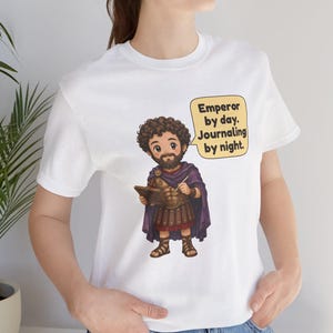 K&ouml;nnte beinhalten: Ein wei&szlig;es T-Shirt mit einer Cartoon-Illustration eines b&auml;rtigen Mannes in r&ouml;mischer R&uuml;stung und violettem Umhang, der ein Buch und einen Stift h&auml;lt. Eine Sprechblase neben der Figur enth&auml;lt den Text: Emperor by day. Journaling by night.