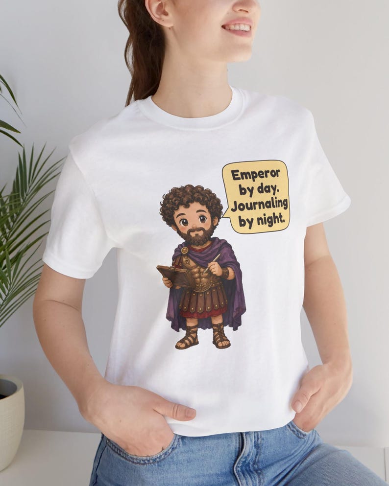 K&ouml;nnte beinhalten: Ein wei&szlig;es T-Shirt mit einer Cartoon-Illustration eines b&auml;rtigen Mannes in r&ouml;mischer R&uuml;stung und violettem Umhang, der ein Buch und einen Stift h&auml;lt. Eine Sprechblase neben der Figur enth&auml;lt den Text: Emperor by day. Journaling by night.
