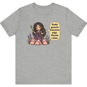 May include: A heather gray t-shirt featuring a cartoon illustration of a dark-haired figure in a purple robe holding a wand and goblet, surrounded by three small pigs. A speech bubble contains the text: Rude guests become pigs. House rules.