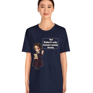 Puede incluir: Una camiseta azul marino con una ilustraci&oacute;n de estilo dibujo animado de una mujer con un vestido oscuro sosteniendo un libro. Un globo de texto contiene el texto blanco: 'Not 'Robert's wife.' Concert pianist, thanks.' El dise&ntilde;o est&aacute; centrado.