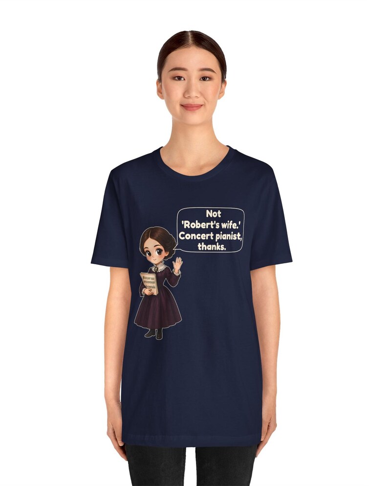 Puede incluir: Una camiseta azul marino con una ilustraci&oacute;n de estilo dibujo animado de una mujer con un vestido oscuro sosteniendo un libro. Un globo de texto contiene el texto blanco: 'Not 'Robert's wife.' Concert pianist, thanks.' El dise&ntilde;o est&aacute; centrado.