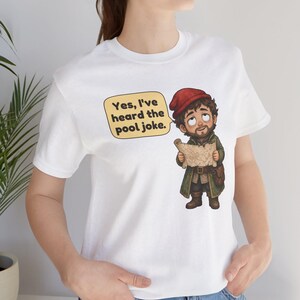 May include: A white t-shirt featuring a cartoon illustration of a bearded character wearing a red cap and green coat, holding a map. A yellow speech bubble next to the character contains the black text, Yes, I've heard the pool joke.