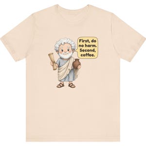 May include: A light beige t-shirt featuring a cartoon illustration of a white-haired, bearded figure in a blue and white toga holding a scroll and a brown pot. A yellow speech bubble contains the text: First, do no harm. Second, coffee.