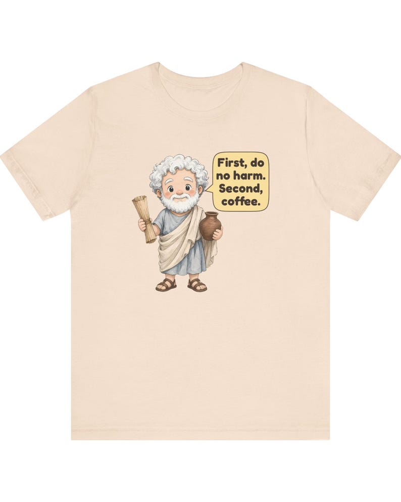 May include: A light beige t-shirt featuring a cartoon illustration of a white-haired, bearded figure in a blue and white toga holding a scroll and a brown pot. A yellow speech bubble contains the text: First, do no harm. Second, coffee.