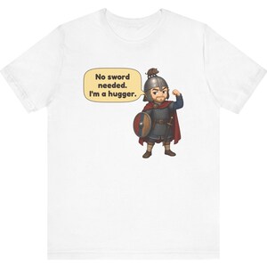 May include: A white t-shirt featuring a cartoon illustration of a warrior in chainmail and a red cape, wearing a helmet with a small animal on top. The warrior holds a round shield and has a speech bubble that says, No sword needed. I'm a hugger.
