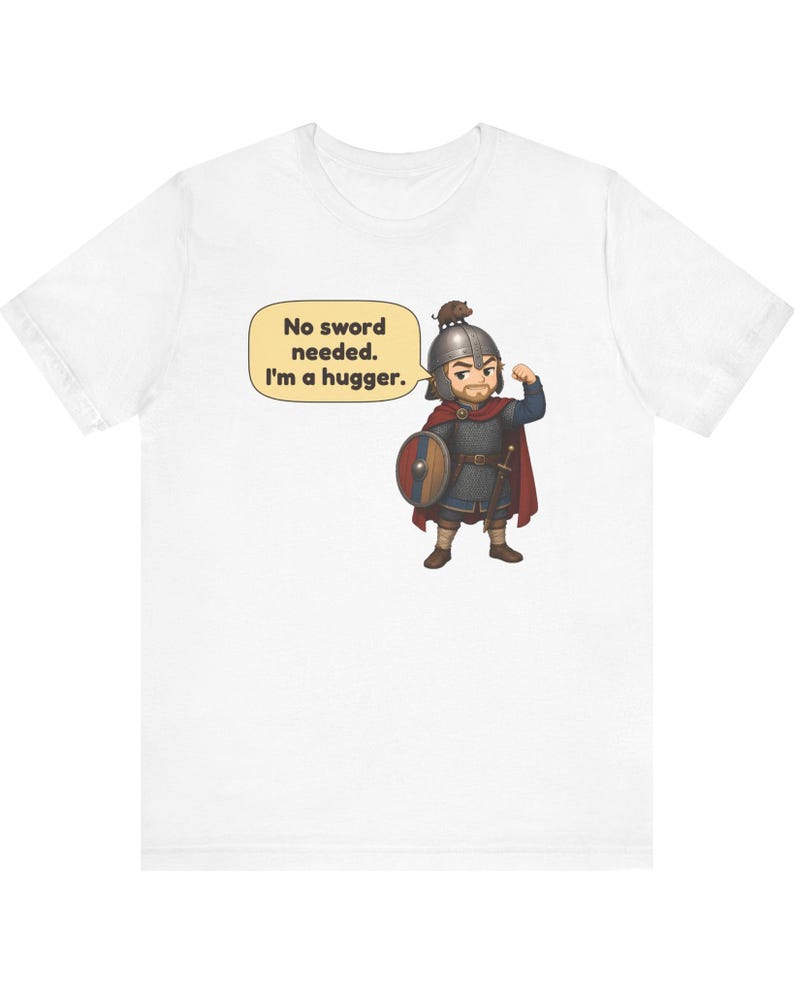 May include: A white t-shirt featuring a cartoon illustration of a warrior in chainmail and a red cape, wearing a helmet with a small animal on top. The warrior holds a round shield and has a speech bubble that says, No sword needed. I'm a hugger.