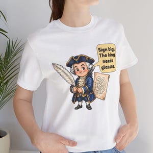 May include: A white t-shirt featuring a cartoon illustration of a historical figure in a blue coat and tricorn hat holding a large white quill. Next to the figure is a speech bubble with the text: Sign big. The king needs glasses.