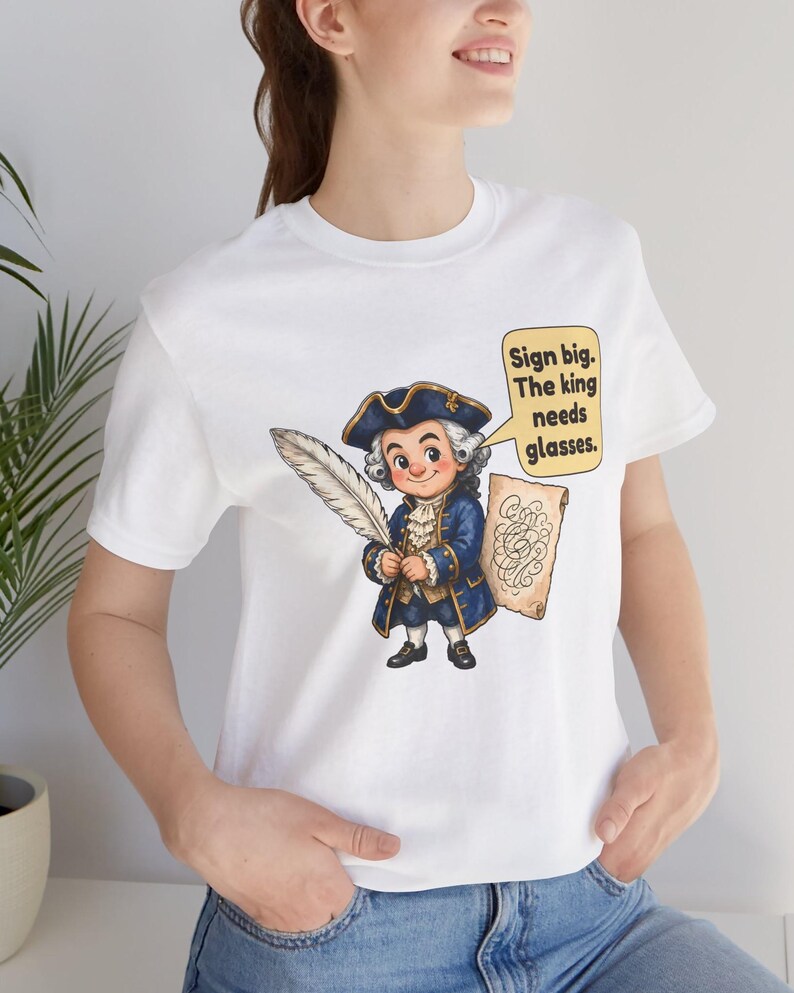 May include: A white t-shirt featuring a cartoon illustration of a historical figure in a blue coat and tricorn hat holding a large white quill. Next to the figure is a speech bubble with the text: Sign big. The king needs glasses.