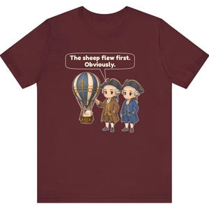 May include: A maroon t-shirt featuring a graphic of two historical figures in tricorn hats standing next to a hot air balloon with a sheep in the basket. A speech bubble above them contains the text: The sheep flew first. Obviously.