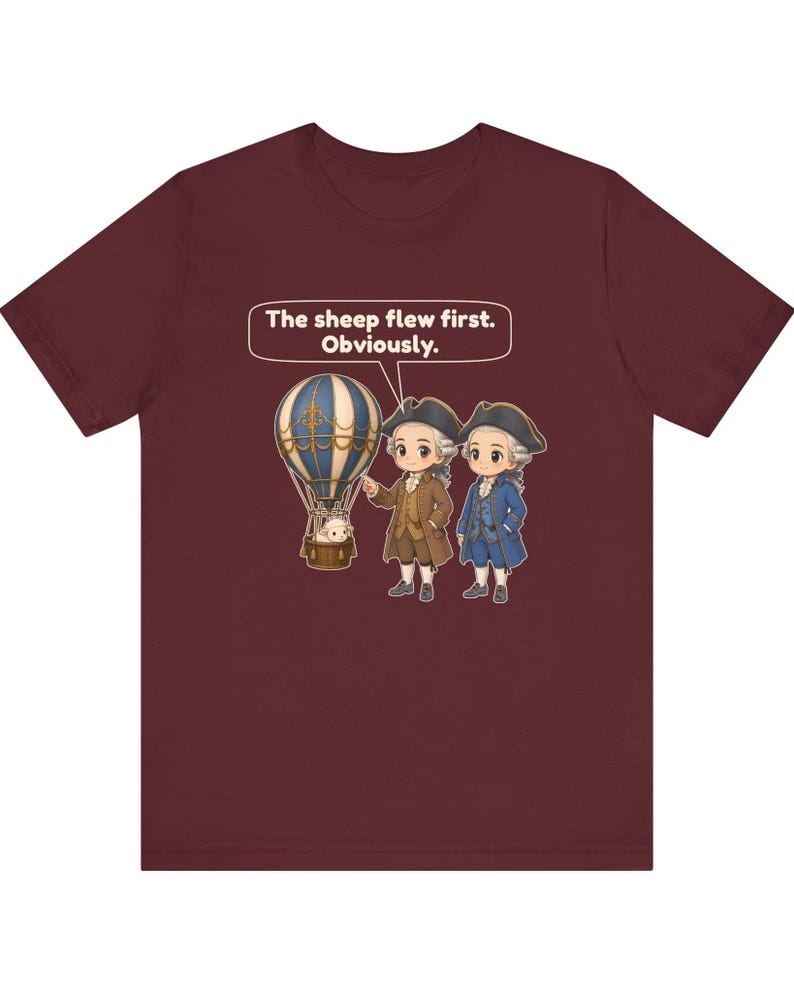May include: A maroon t-shirt featuring a graphic of two historical figures in tricorn hats standing next to a hot air balloon with a sheep in the basket. A speech bubble above them contains the text: The sheep flew first. Obviously.