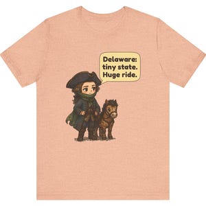 May include: A peach-colored t-shirt featuring a cartoon illustration of a person in 18th-century attire standing next to a small horse. A speech bubble contains the text: Delaware: tiny state. Huge ride.