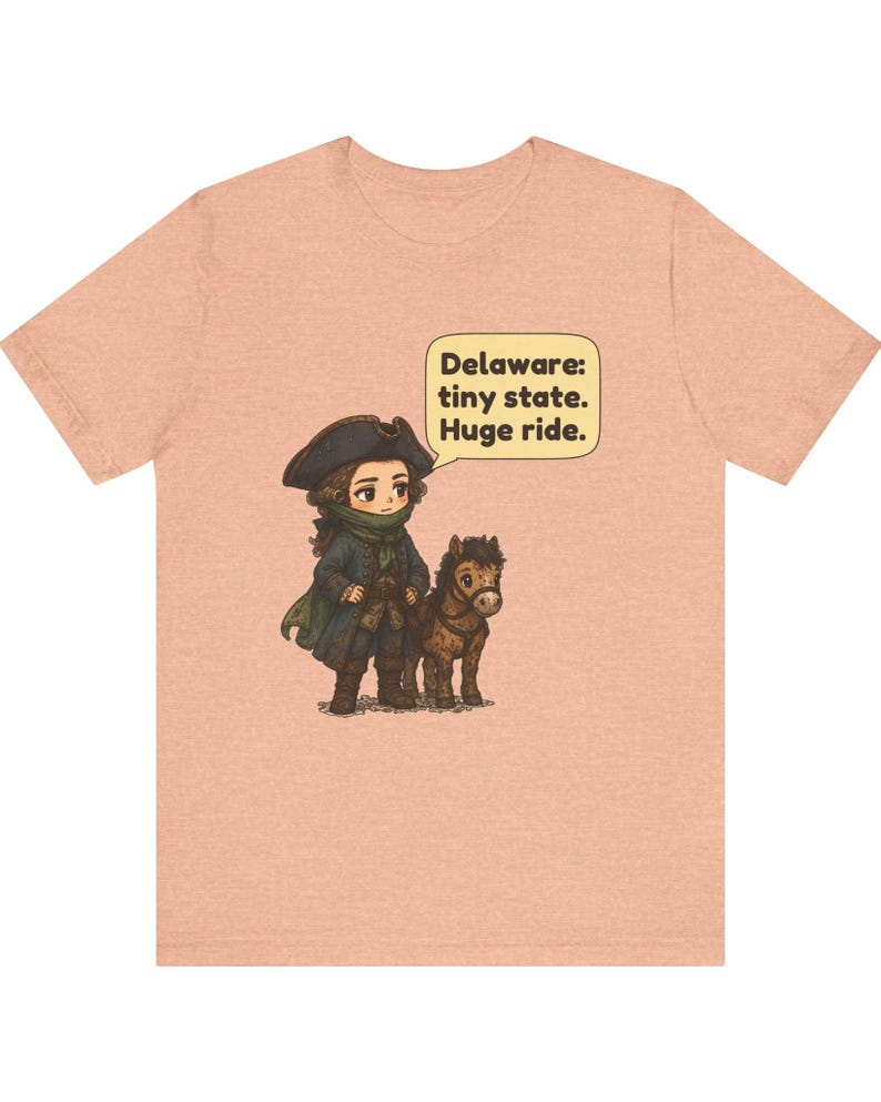 May include: A peach-colored t-shirt featuring a cartoon illustration of a person in 18th-century attire standing next to a small horse. A speech bubble contains the text: Delaware: tiny state. Huge ride.