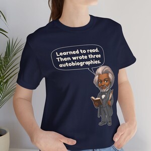 Puede incluir: Una camiseta azul marino con una ilustraci&oacute;n de dibujos animados de Frederick Douglass sosteniendo un libro y una pluma. Un globo de texto blanco sobre la figura contiene el texto: Learned to read. Then wrote three autobiographies.