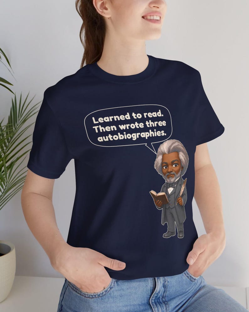 Puede incluir: Una camiseta azul marino con una ilustraci&oacute;n de dibujos animados de Frederick Douglass sosteniendo un libro y una pluma. Un globo de texto blanco sobre la figura contiene el texto: Learned to read. Then wrote three autobiographies.