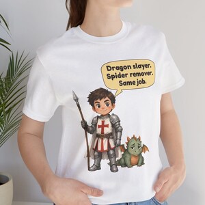 K&ouml;nnte beinhalten: Ein wei&szlig;es T-Shirt mit einer Grafik eines Cartoon-Ritters in silberner R&uuml;stung, der einen Speer h&auml;lt und neben einem kleinen gr&uuml;nen Drachen steht. Eine gelbe Sprechblase dar&uuml;ber enth&auml;lt den Text: Dragon slayer. Spider remover. Same job.