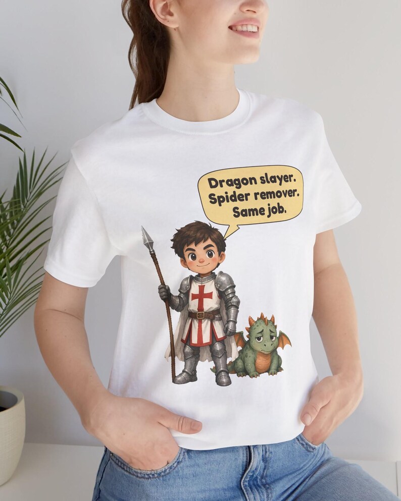 K&ouml;nnte beinhalten: Ein wei&szlig;es T-Shirt mit einer Grafik eines Cartoon-Ritters in silberner R&uuml;stung, der einen Speer h&auml;lt und neben einem kleinen gr&uuml;nen Drachen steht. Eine gelbe Sprechblase dar&uuml;ber enth&auml;lt den Text: Dragon slayer. Spider remover. Same job.