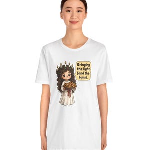 May include: A white t-shirt featuring a cartoon illustration of a figure wearing a crown of candles and a white robe, holding a tray of buns. A speech bubble next to the figure contains the text: Bringing the light (and the buns).