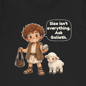 May include: A black t-shirt featuring a cartoon illustration of a curly-haired person in a fur vest holding a sling and a stone, standing next to a white lamb. A speech bubble contains the text: Size isn't everything. Ask Goliath.