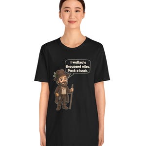 May include: A black short-sleeve t-shirt featuring a cartoon illustration of a bearded hiker wearing a brown hat and coat, holding a walking stick. A speech bubble above the hiker contains the text: I walked a thousand miles. Pack a lunch.