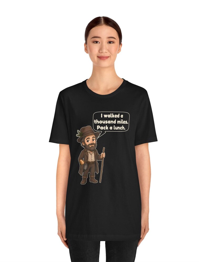 May include: A black short-sleeve t-shirt featuring a cartoon illustration of a bearded hiker wearing a brown hat and coat, holding a walking stick. A speech bubble above the hiker contains the text: I walked a thousand miles. Pack a lunch.