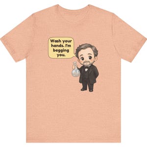 May include: A peach-colored t-shirt featuring a cartoon illustration of a man in a black suit and bowtie holding a glass flask. A speech bubble next to him contains the text: Wash your hands. I'm begging you.