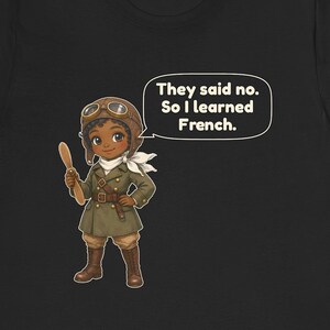May include: A black t-shirt featuring a cartoon illustration of a pilot wearing a brown leather aviator cap, goggles, and a green coat. The character holds a wooden propeller and has a speech bubble that says, "They said no. So I learned French."