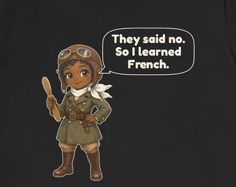 May include: A black t-shirt featuring a cartoon illustration of a pilot wearing a brown leather aviator cap, goggles, and a green coat. The character holds a wooden propeller and has a speech bubble that says, "They said no. So I learned French."