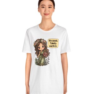 May include: A white t-shirt featuring a graphic of a cartoon character with long brown hair, a flower crown, and a green dress, holding a pomegranate. A yellow speech bubble next to the character contains the text: Six seeds. Totally worth it.